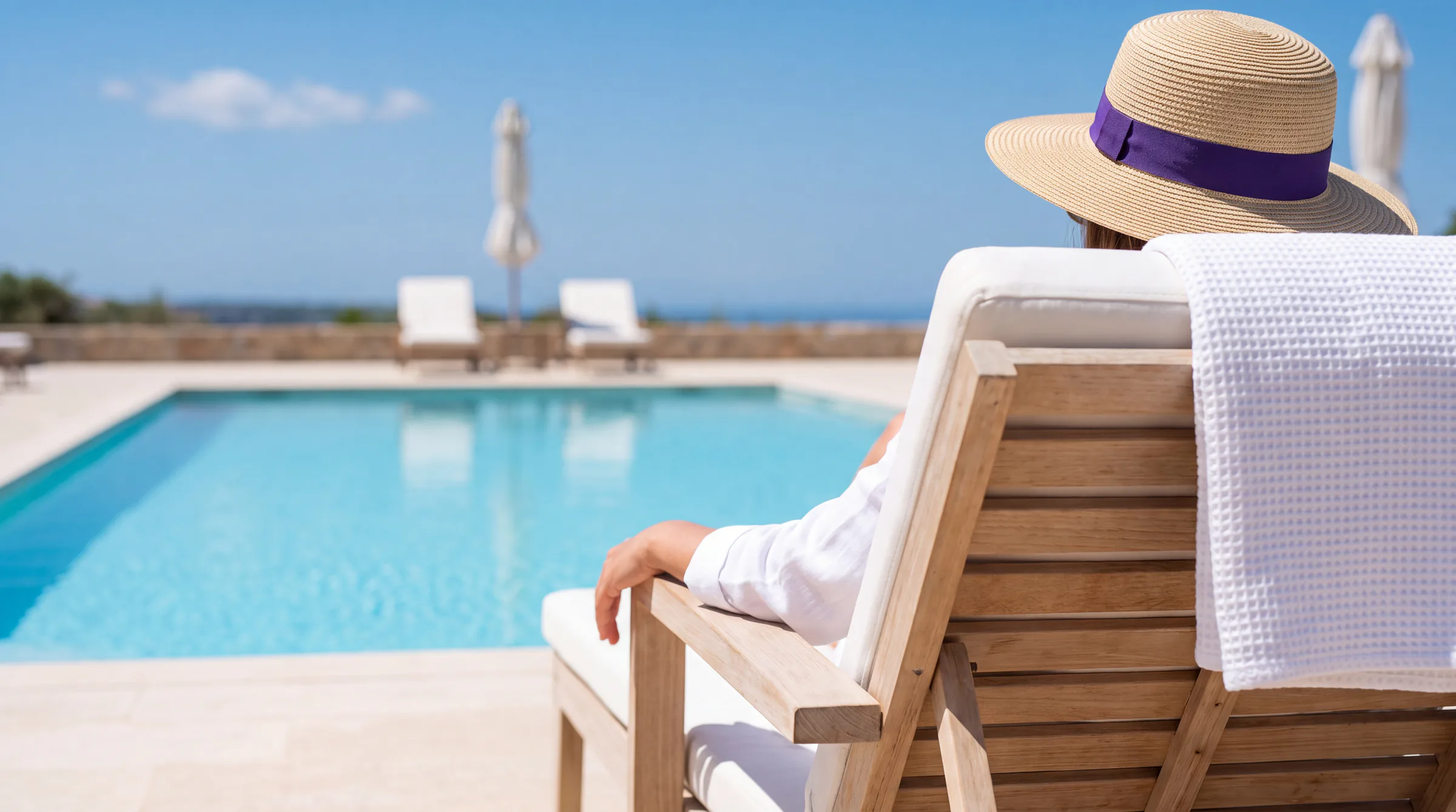 A hotel guest in a wide straw hat with a violet ribbon, resting on a lounger beside a pool